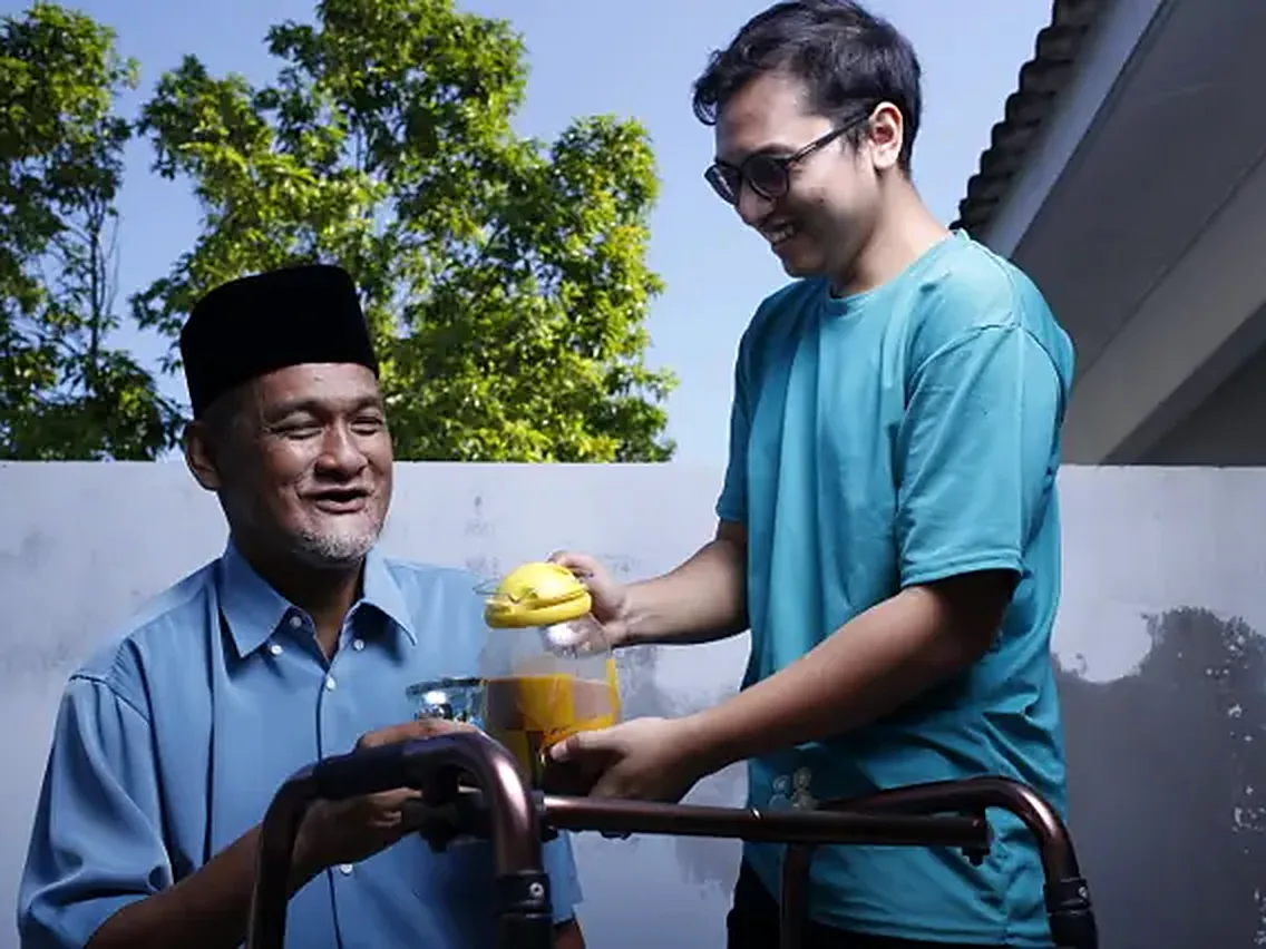 A smiling young caregiver helps a senior man at his walking frame, outdoors in the sun